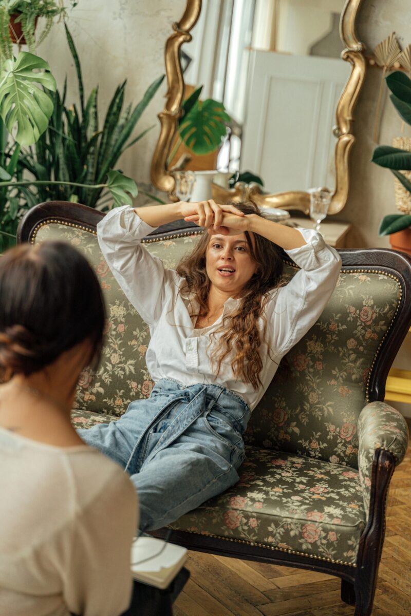 Two women engage in conversation on a vintage floral sofa, surrounded by plants and classic decor.
